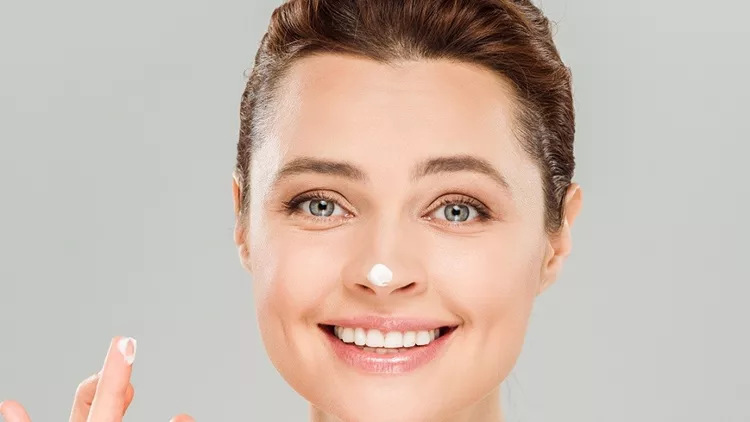 cheerful woman applying face cream and smiling isolated on grey