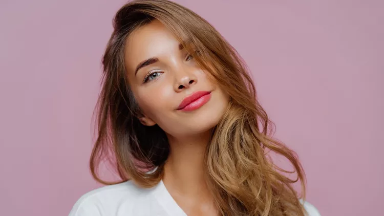 Portrait of satisfied relaxed young female model tilts head, has makeup, fair hair, dressed in white clothes, poses against purple background, has well cared complexion. People, beauty, face care