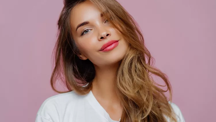 Portrait of satisfied relaxed young female model tilts head, has makeup, fair hair, dressed in white clothes, poses against purple background, has well cared complexion. People, beauty, face care