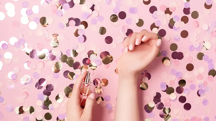 Luxury perfume concept. Female hands holding stylish bottle of perfume on pastel pink background with colorful round paper confetti. Creative trendy flat lay with space for text.