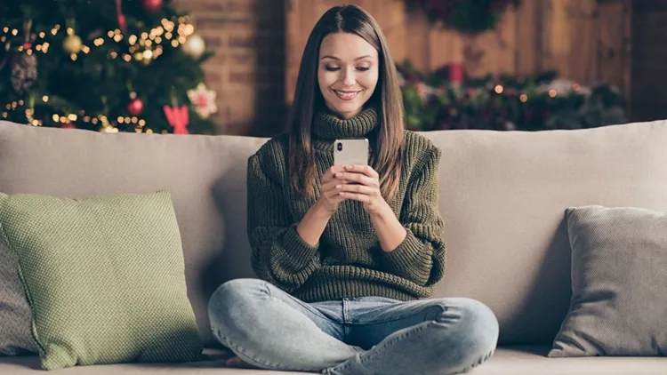 Full body photo of positive festive girl sit on divan crossing her legs using cellphone communicating enjoy christmas celebration in house full of newyear decoration lights indoors