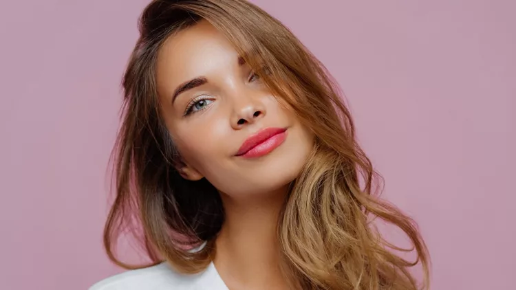 Portrait of satisfied relaxed young female model tilts head, has makeup, fair hair, dressed in white clothes, poses against purple background, has well cared complexion. People, beauty, face care