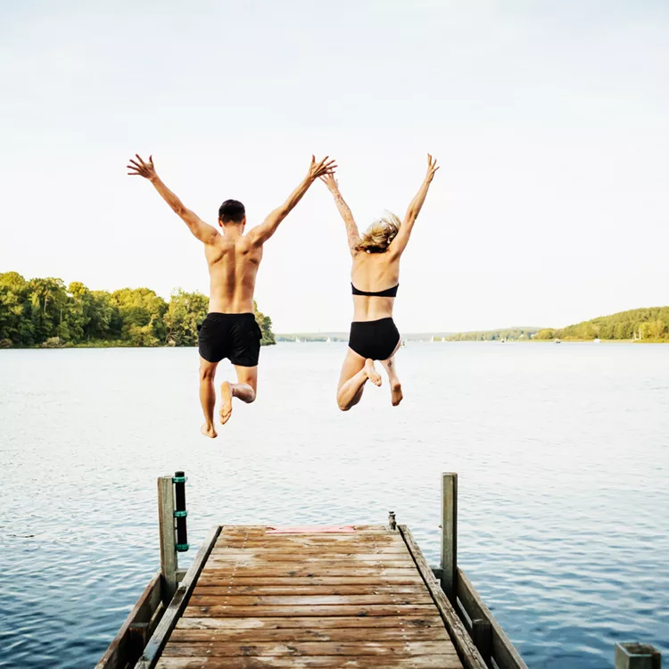 Two Friends Jumping Off Jetty At Lake Together